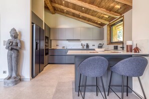 A modern kitchen with a stone Buddha statue, a stainless steel refrigerator, and a large island with bar stools. - Newly Renovated 4-Bedroom Chalet - Mont Blanc Views, Hot Tub & Sauna (Chamonix-Mont-Blanc)