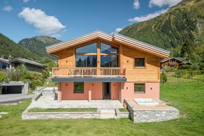 A two-story wooden cabin with a balcony, stone foundation, and a hot tub in the front yard. - Newly Renovated 4-Bedroom Chalet - Mont Blanc Views, Hot Tub & Sauna (Chamonix-Mont-Blanc)