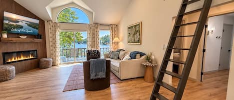 A spacious living room with a fireplace, a television, and a large window overlooking a lake.