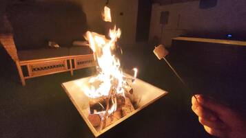 Un pozo de fuego cuadrado con llamas, un banco de madera y una mesa con un malvavisco en un palo.