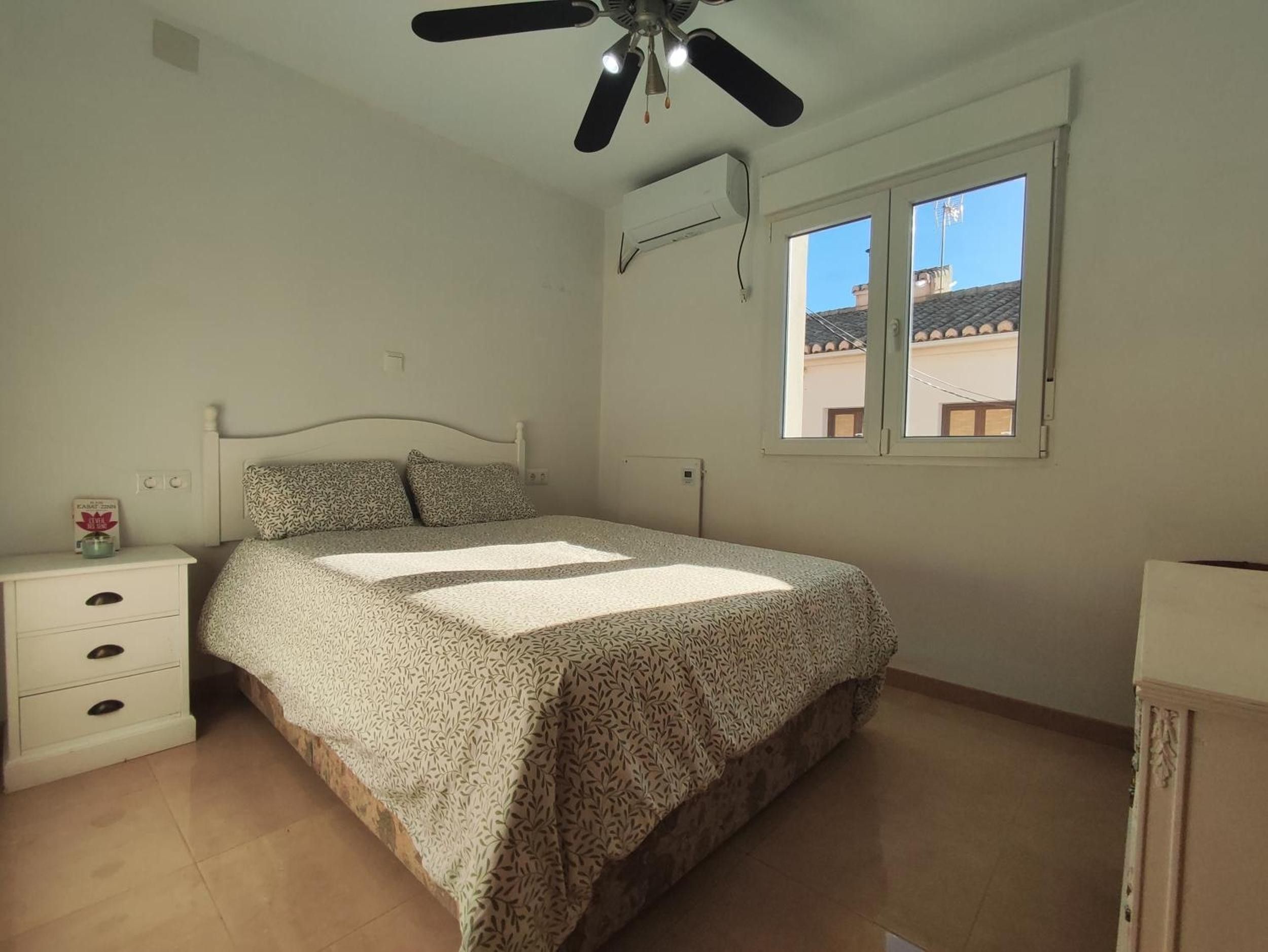A bedroom with a bed, a nightstand, a ceiling fan, and a window with a view of buildings.