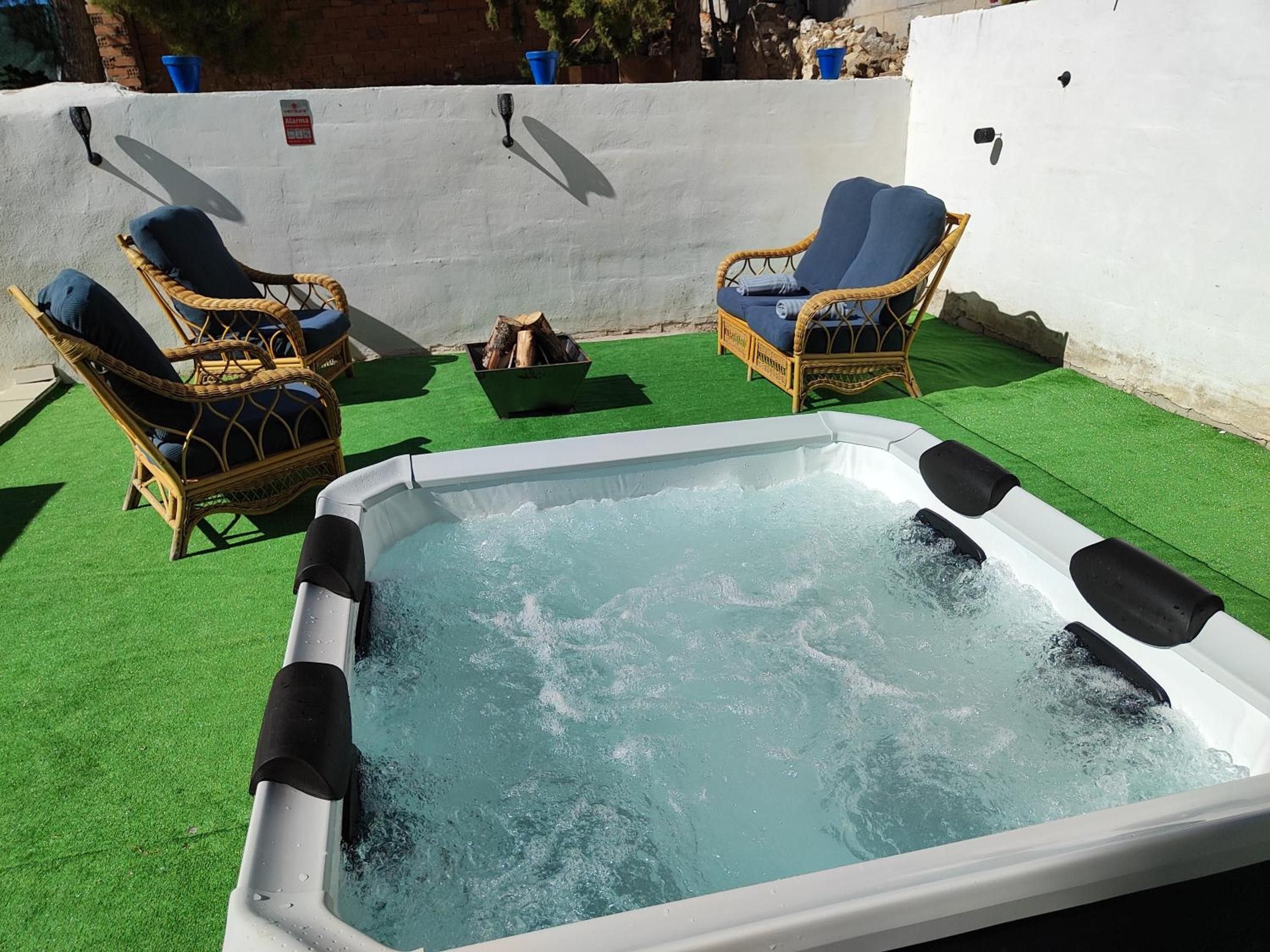 A hot tub with two wicker chairs and a fire pit on a green artificial lawn.