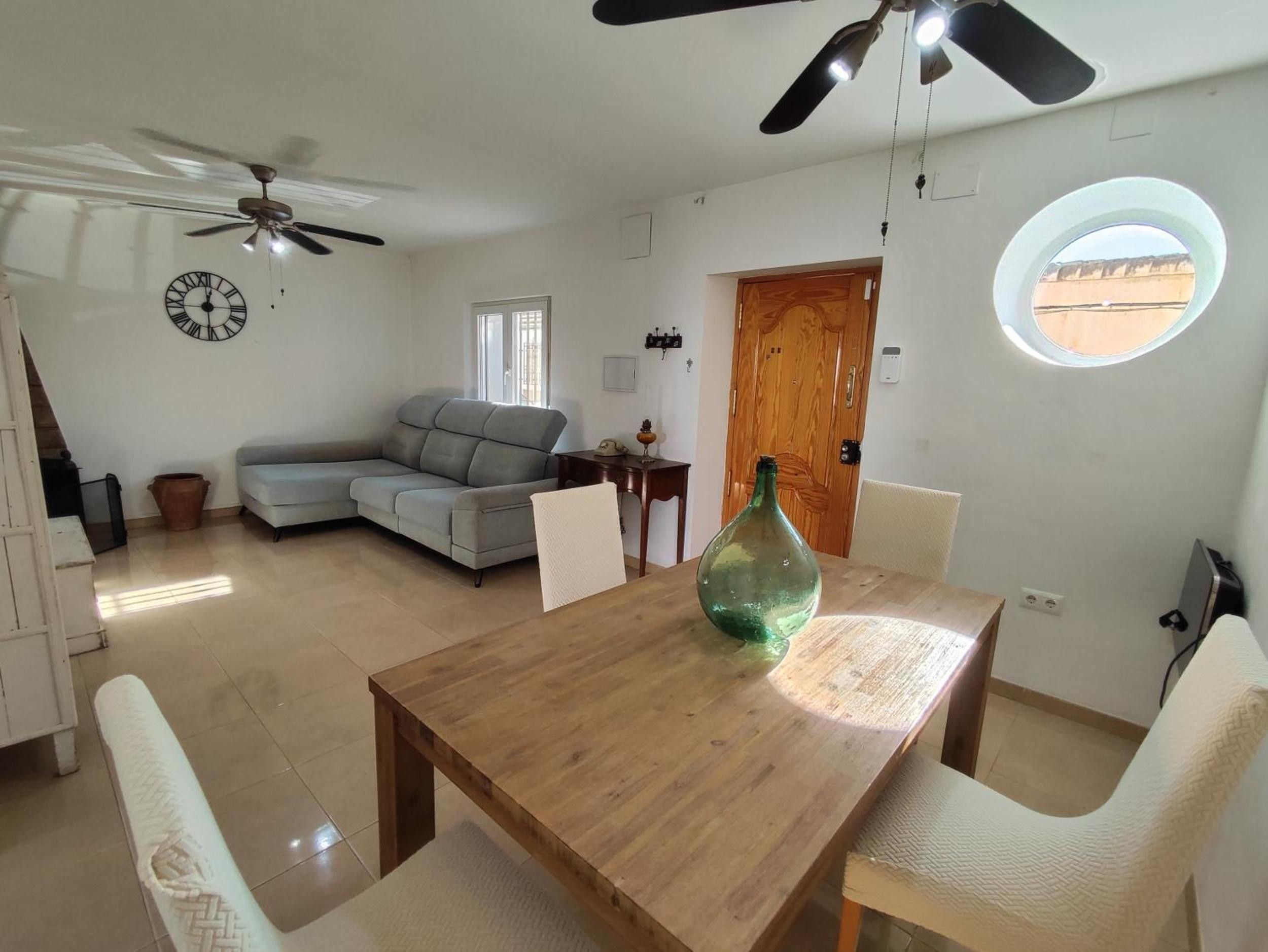 A living room with a sofa, a wooden dining table with chairs, a ceiling fan, a clock, and a window with a view.