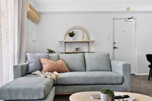 A modern living room with a grey sectional sofa, a small round coffee table, and a white door. - Coastal Views – Mosman Park – 2BR with Balcony & Pool (Mosman Park)