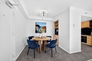 A dining area with a round table, four chairs, a painting, and a kitchen in the background. - Coastal Views – Mosman Park – 2BR with Balcony & Pool (Mosman Park)