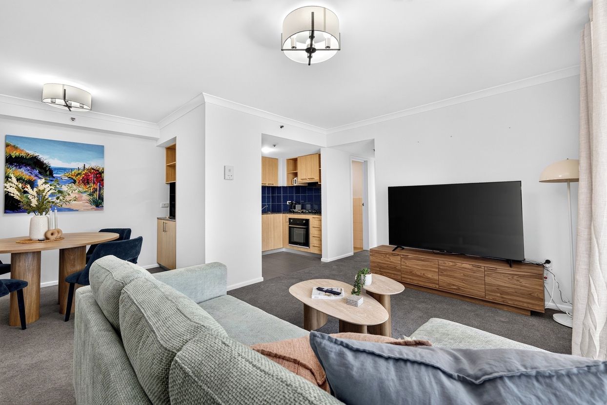 A modern living room with a sofa, coffee table, and a flat-screen TV. There is a dining area with a table and chairs, and a kitchen with cabinets and a tiled backsplash.