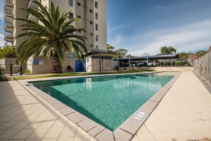 A swimming pool with a clear blue water, surrounded by a tiled deck, a palm tree, and a covered seating area. - Wellington Breeze – Mosman Park – 2BR with Pool & Balcony (Mosman Park)