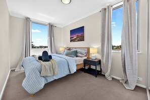 A bedroom with a bed, bedside tables, a window with curtains, and a painting on the wall. - Wellington Breeze – Mosman Park – 2BR with Pool & Balcony (Mosman Park)