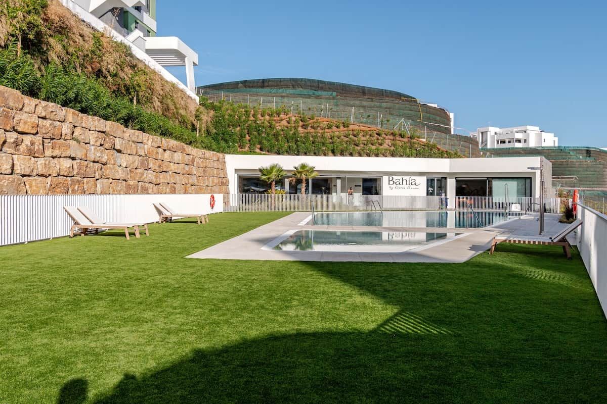 A modern building with a pool, lounge chairs, and a view of a hill.
