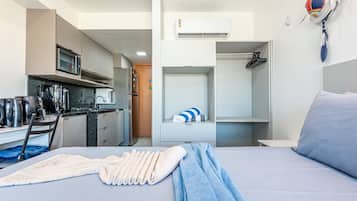 Pieni hotellihuone, jossa on minikeittiö, sinivalkoraidallinen tyyny sängyllä ja valkoinen ilmastointilaite seinällä.