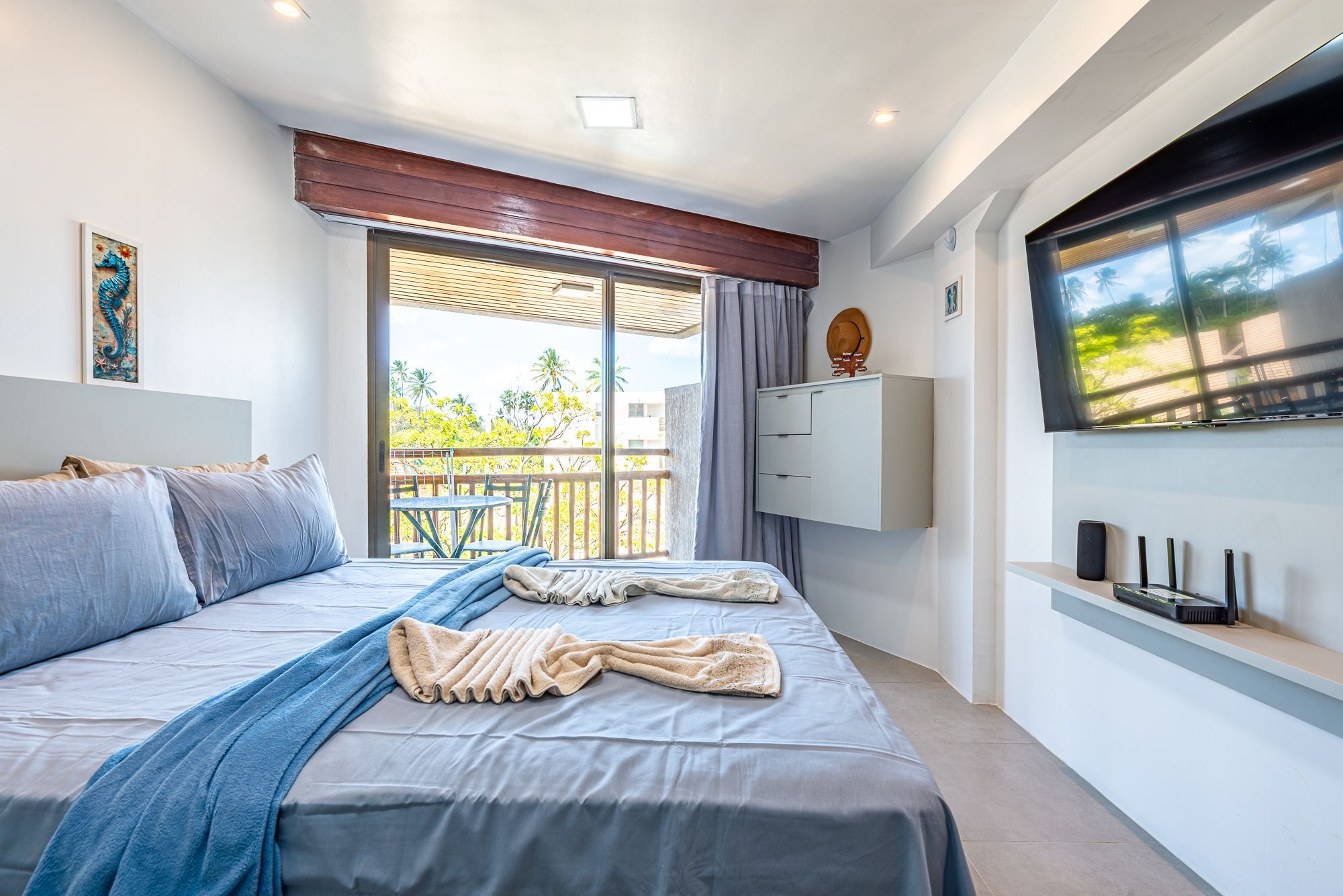 A modern bedroom with a bed, a TV, a balcony, and a view of palm trees.