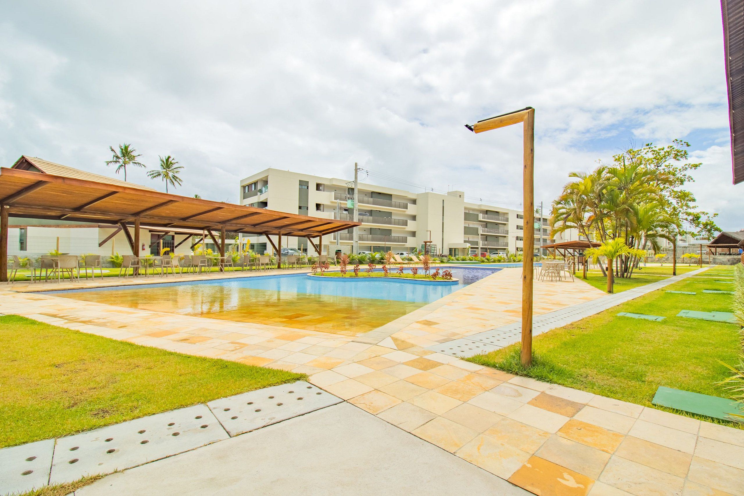 A swimming pool with a tiled deck, a covered seating area, and a building in the background.