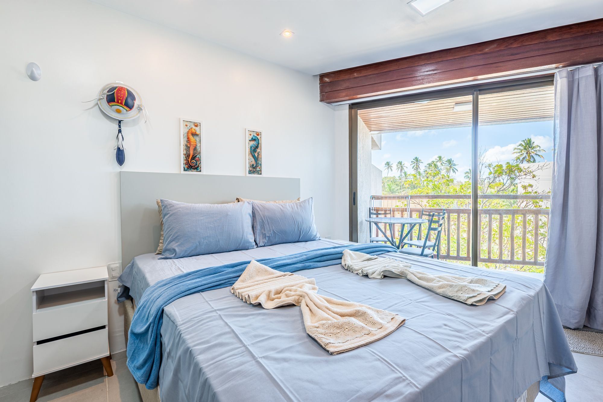 A bedroom with a bed, a nightstand, and a window with a view of palm trees.