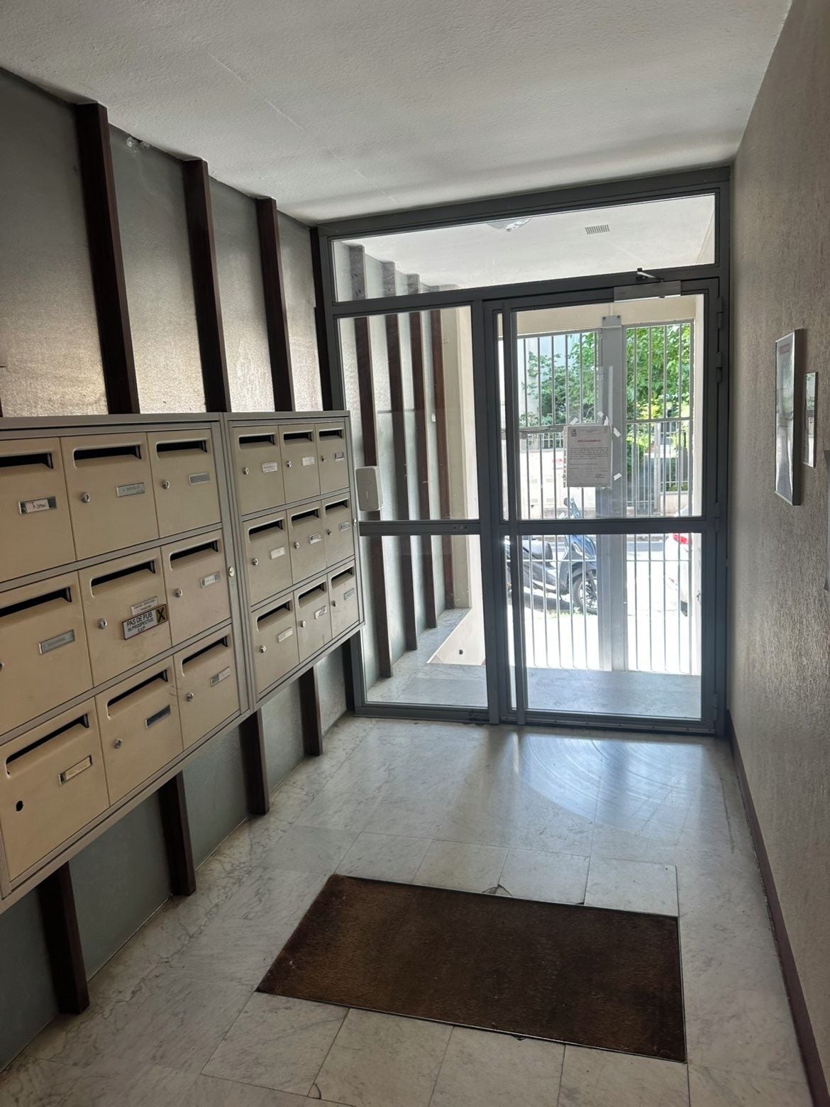 A hallway with a metal mail slot system, a sliding glass door leading outside, and a brown mat at the entrance.