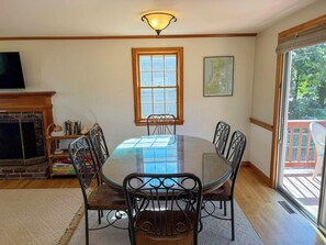 A dining area with a glass-top table, metal chairs, a fireplace, a bookshelf, and a window with a view. - Relax and enjoy all that the Cape has to offer in this 3 bedroom home. (Dennis Port)