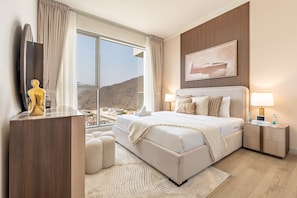 A hotel room with a large bed, bedside lamps, a nightstand, a wooden dresser, a large window with a view of a desert landscape, and a framed picture on the wall. - Majestic Hajar Mountain Views 1BR and Beach Access, Near Khorfakkan Amphitheatre (Khor Fakkan)