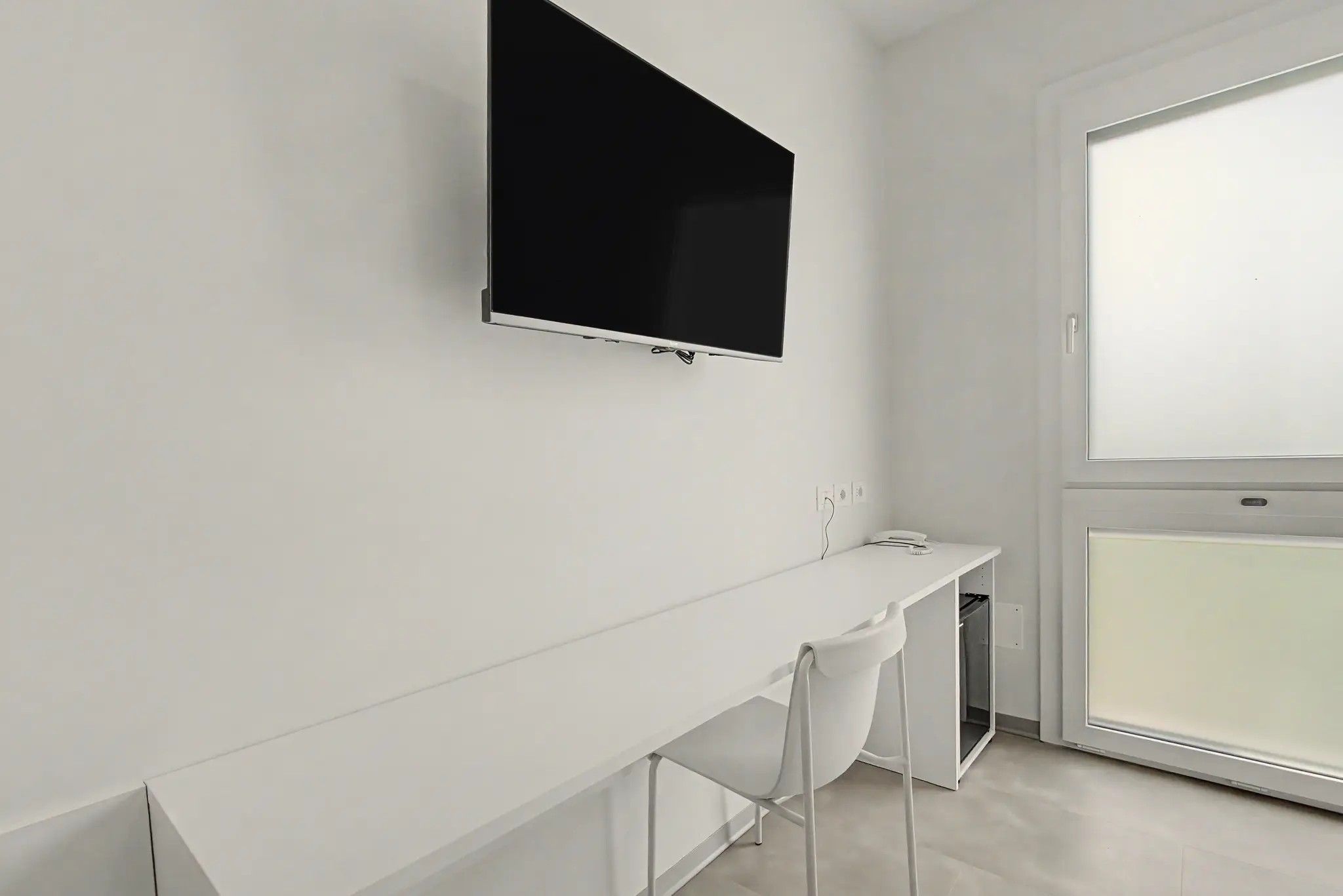 A modern, minimalistic room with a flat-screen TV mounted on the wall, a white desk with a chair, and a large window providing natural light.