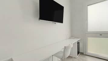 A modern, minimalistic room with a flat-screen TV mounted on the wall, a white desk with a chair, and a large window providing natural light.
