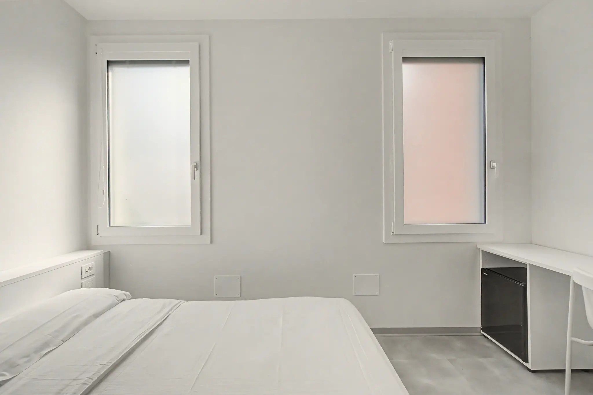 Ein schlicht eingerichtetes Schlafzimmer mit einem Bett, zwei Fenstern, einem Nachttisch und einem Einbauschrank.