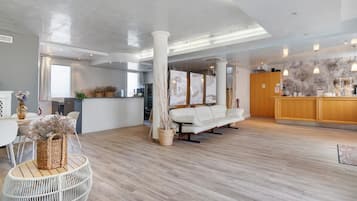 A spacious hotel lobby with a wooden floor, a white sofa, a reception desk, and a large window.