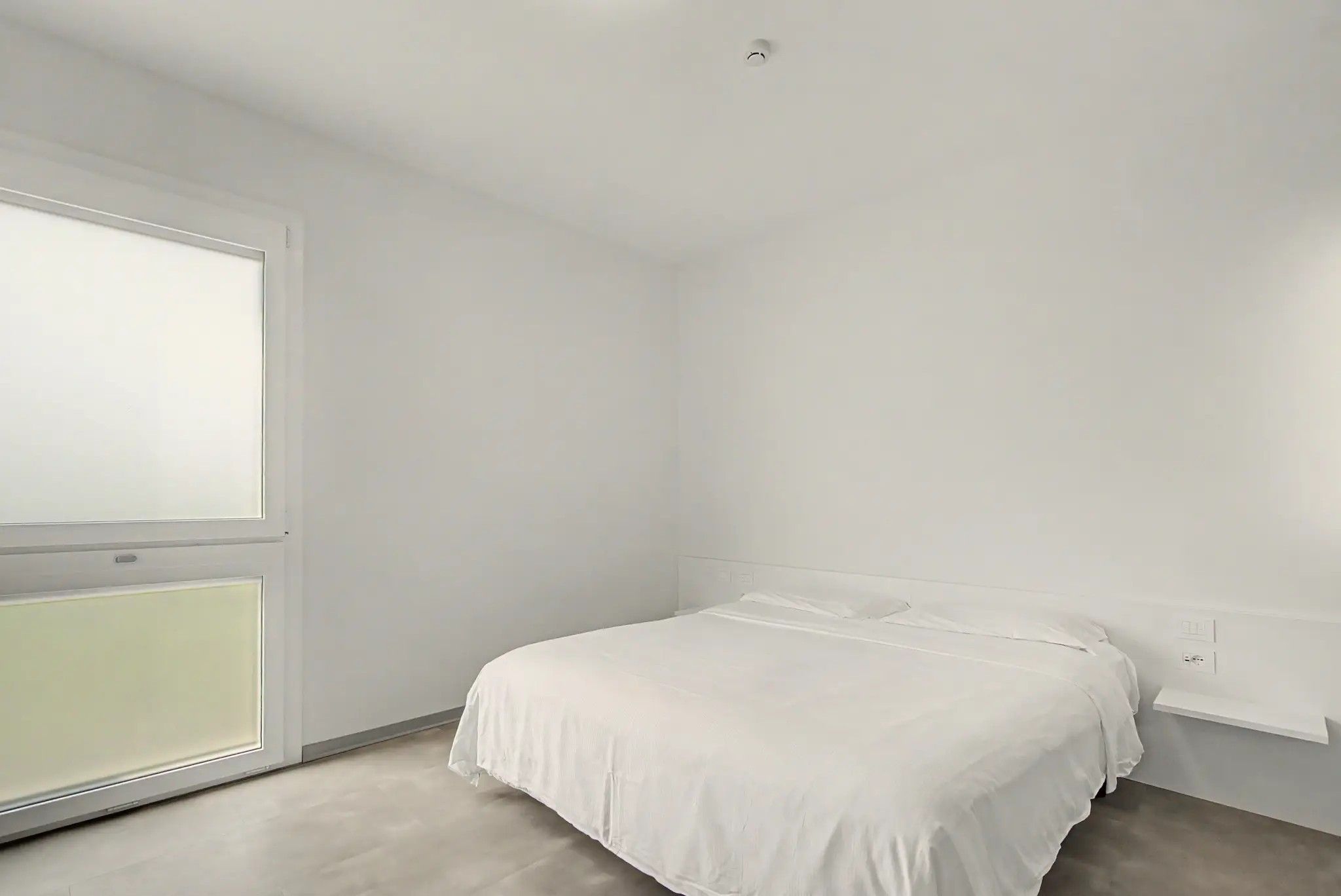 Une chambre à coucher minimaliste avec un lit simple, une fenêtre et un mur blanc.