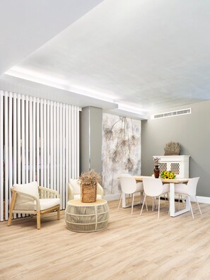 Una sala de estar moderna con comedor, que incluye un sofá blanco, mobiliario de mimbre y una pared decorativa con un mural floral.
