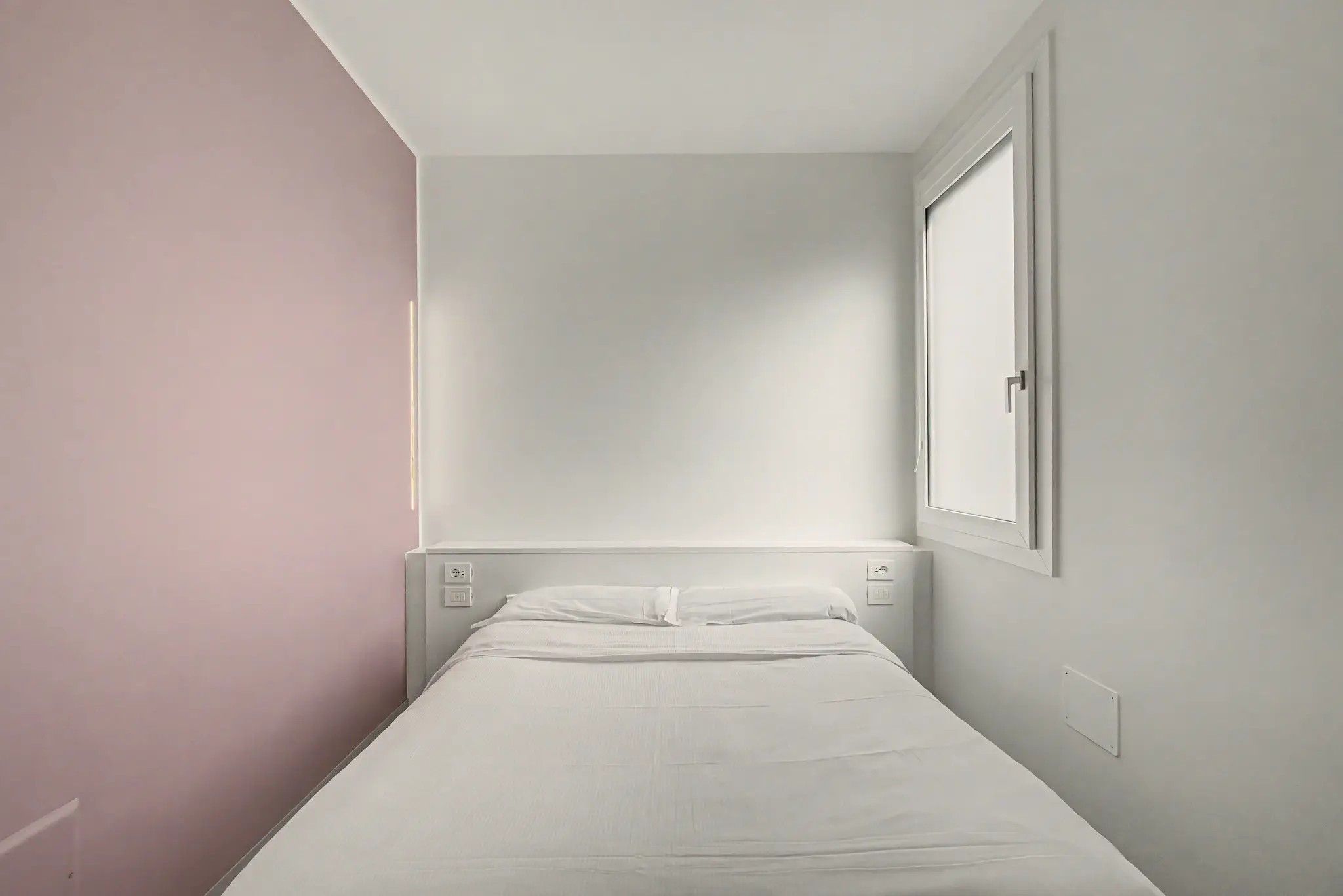 A small, single-bed room with a plain white bed, a nightstand, and a wall-mounted light.