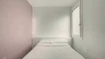 A small, single-bed room with a plain white bed, a nightstand, and a wall-mounted light.