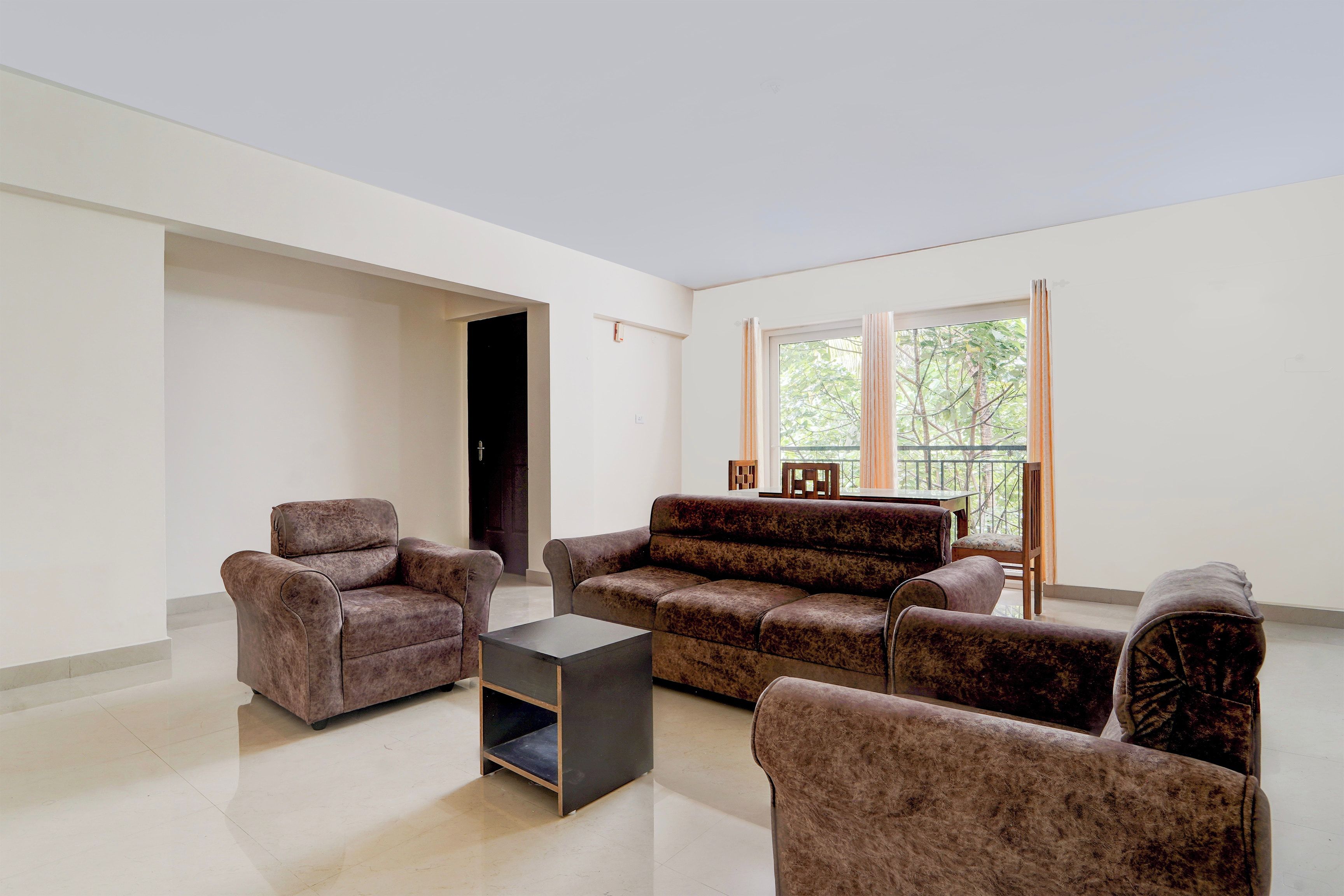 Una sala de estar con un conjunto de sofá, una mesa de centro y una mesa de comedor con sillas. Hay una puerta que conduce a otra habitación y una ventana con cortinas.