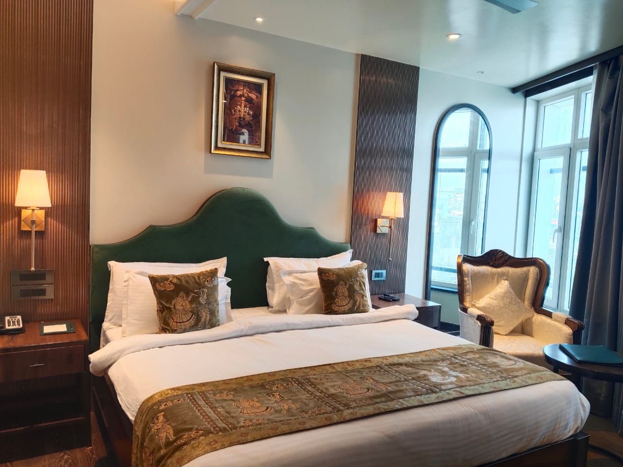 A hotel room with a large bed, two bedside lamps, a headboard, a window with curtains, and a framed picture on the wall.