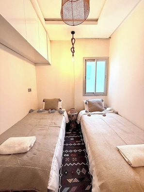 Une petite chambre agréable, dotée de deux lits simples, d’une fenêtre et d’un luminaire suspendu.