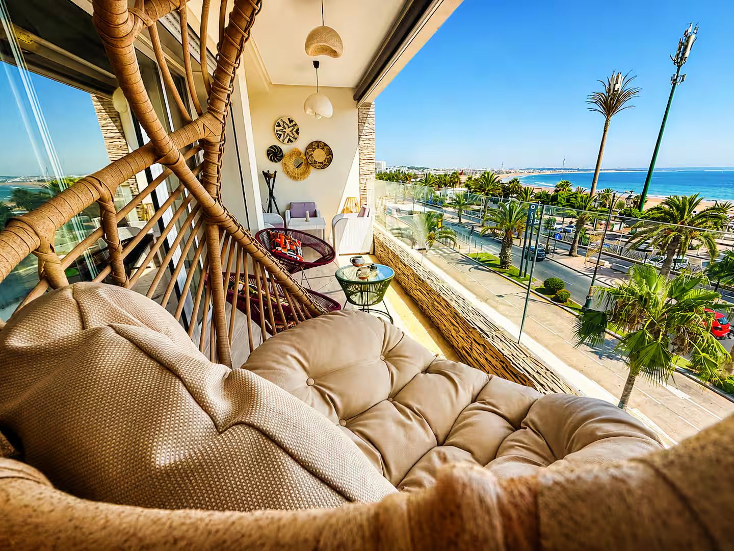 Un balcon avec un fauteuil en osier, un siège rembourré, une petite table et une vue sur la plage et l’océan.