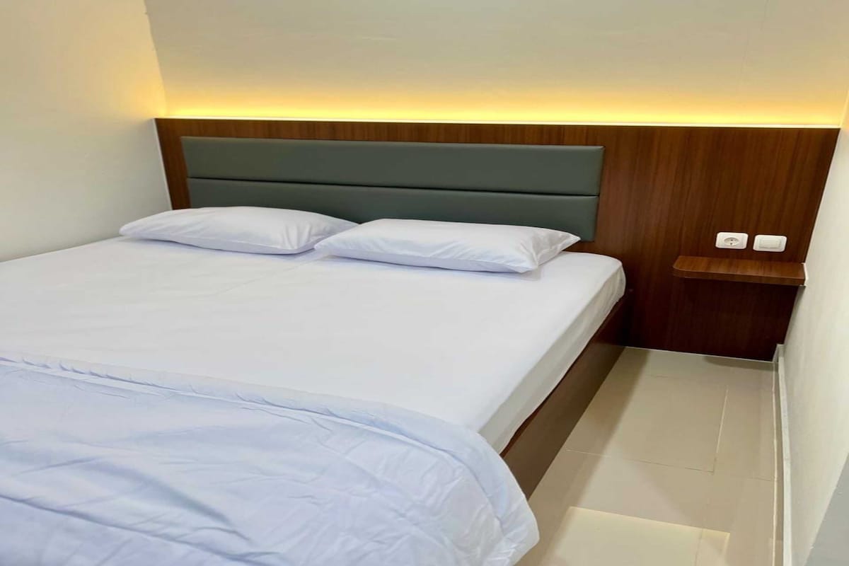 A hotel room with a bed, two pillows, a nightstand with a lamp, and a wooden headboard.