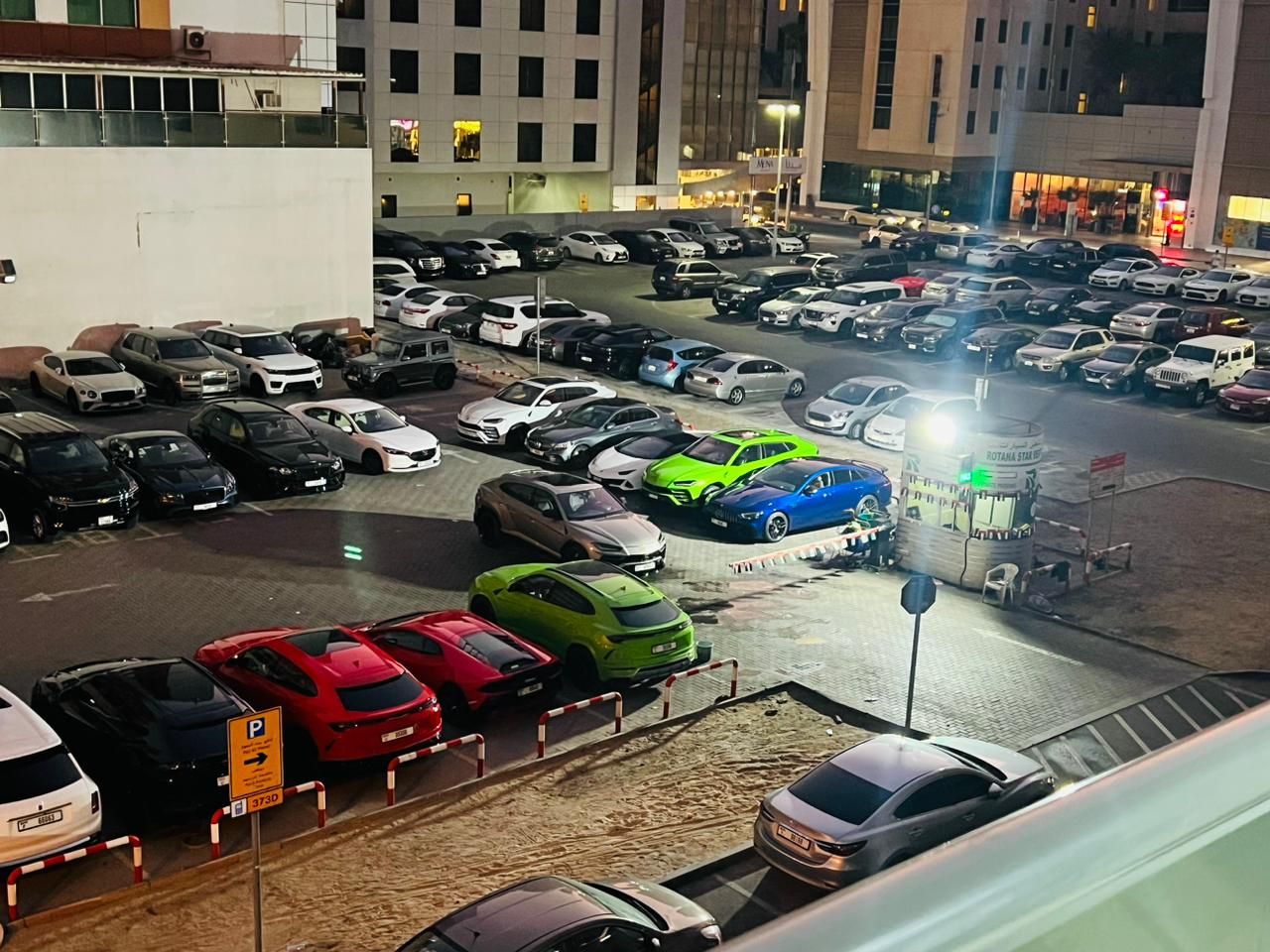 Een parkeerplaats met meerdere auto's, een bord dat een parkeerterrein aangeeft, en een gebouw met verlichte ramen op de achtergrond.