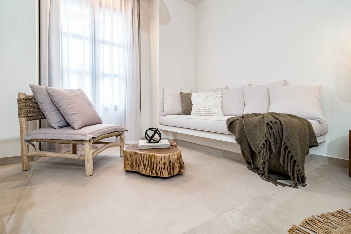 Een moderne woonkamer met een houten stoel, een ronde houten salontafel, een witte bank en een groot raam met dunne gordijnen.