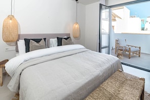 Un dormitorio con una cama, mesitas de noche, un banco y vistas a través de un ventanal.