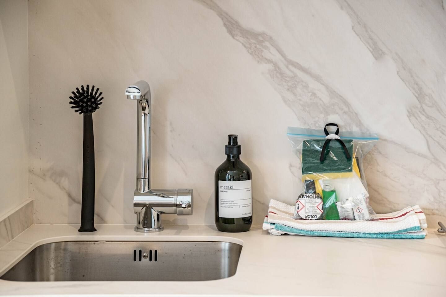 A bathroom sink with a chrome faucet, a black hairbrush, a bottle of Meraki, and a towel with cleaning supplies.