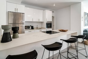 A modern kitchen with white cabinetry, a stainless steel refrigerator, a microwave, an oven, and a black induction cooktop. There are three black bar stools and two decorative vases on the counter. - AB3 - Spacious holiday home, Puerto Banus (Marbella)