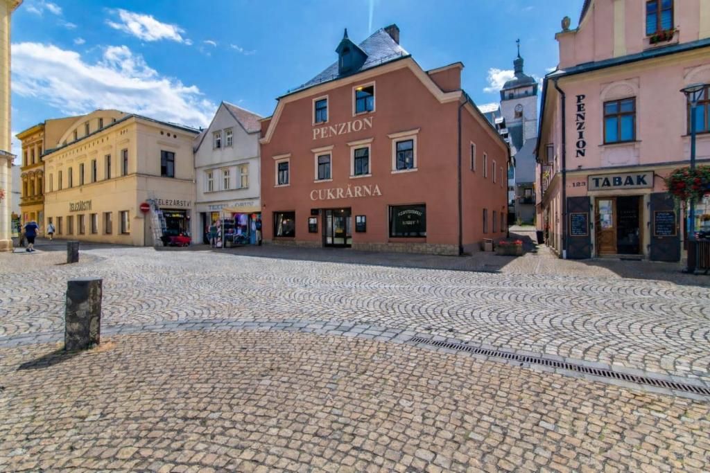 Una plaza adoquinada con edificios históricos, incluyendo un edificio rojo con la inscripción 'PENZION CUKRARNA' y un edificio rosa con la inscripción 'TABAK'.