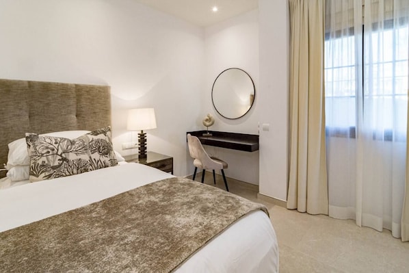 Une chambre d’hôtel avec un lit, un bureau, une chaise, un miroir rond et une fenêtre avec des rideaux.