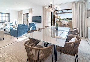 A modern hotel room with a large glass dining table, comfortable seating, a flat-screen TV, and a view of the outdoors. - AB26 - Lovely holiday home in Aldea Blanca (Marbella)