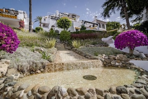Um jardim com um caminho de pedra que leva a uma piscina, rodeado por edifícios brancos e buganvílias roxas.