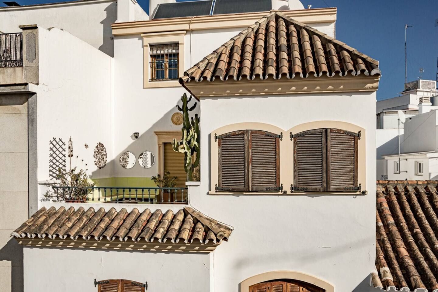 Um edifício com fachada branca, telhas de barro, persianas de madeira e uma varanda com guarda-corpo metálico.