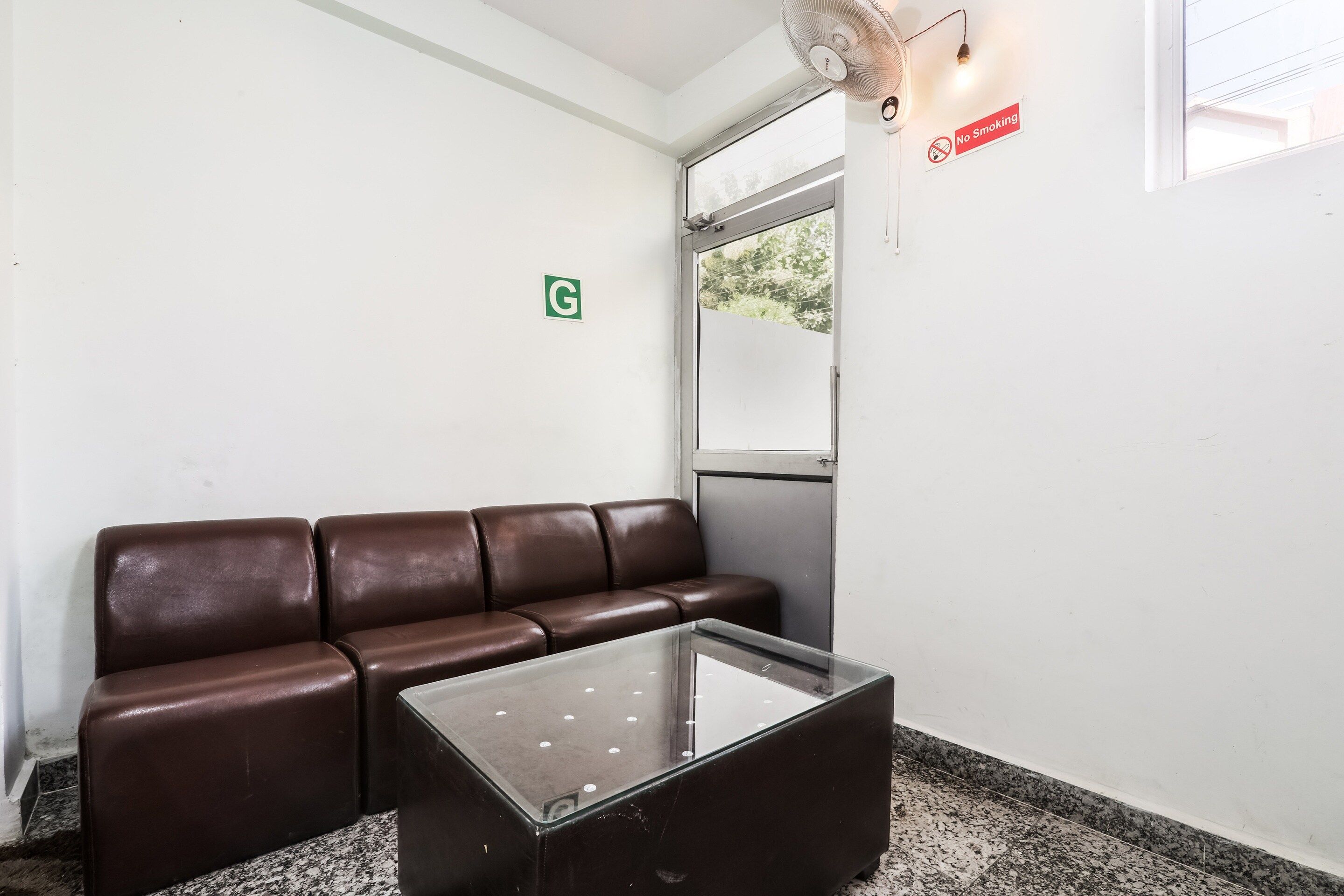 A waiting area with a brown leather couch and a glass-top table, a 'No Smoking' sign, a 'G' restroom sign, and a window with a view of trees.