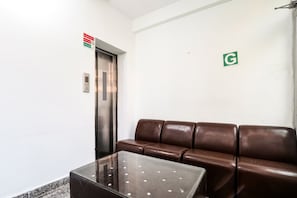A waiting area with a brown leather couch, a glass-top table, and an elevator. - Hotel O Kiara Inn (Lucknow)