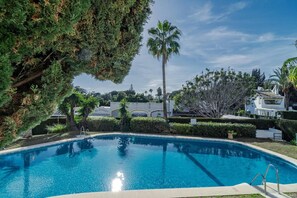 Piscina com superfície de água azul cristalina, cercada por vegetação bem cuidada e um edifício branco ao fundo.
