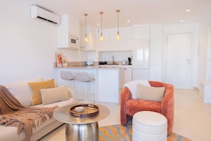 A modern living room with a sofa, ottoman, and a coffee table with a decorative bowl. There is a kitchen area with white cabinets and a bar stool. - AC152 - Top Renovated Modern 1 Bed with Pool (Marbella)