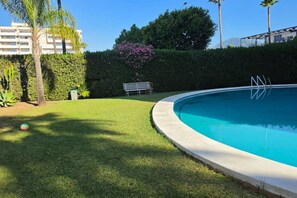 A swimming pool with a grassy area and a bench, surrounded by a hedge and palm trees. - AC152 - Top Renovated Modern 1 Bed with Pool (Marbella)