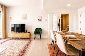 Ein modernes Wohnzimmer mit einem Flachbildfernseher, einem grünen Sessel, einem Esstisch mit Stühlen und einem rot-weißen Teppich.