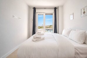 A hotel room with a bed, a set of towels, a window with curtains, and a view of a pool and landscape. - AH2B - Modern apartment near Estepona town (Estepona)